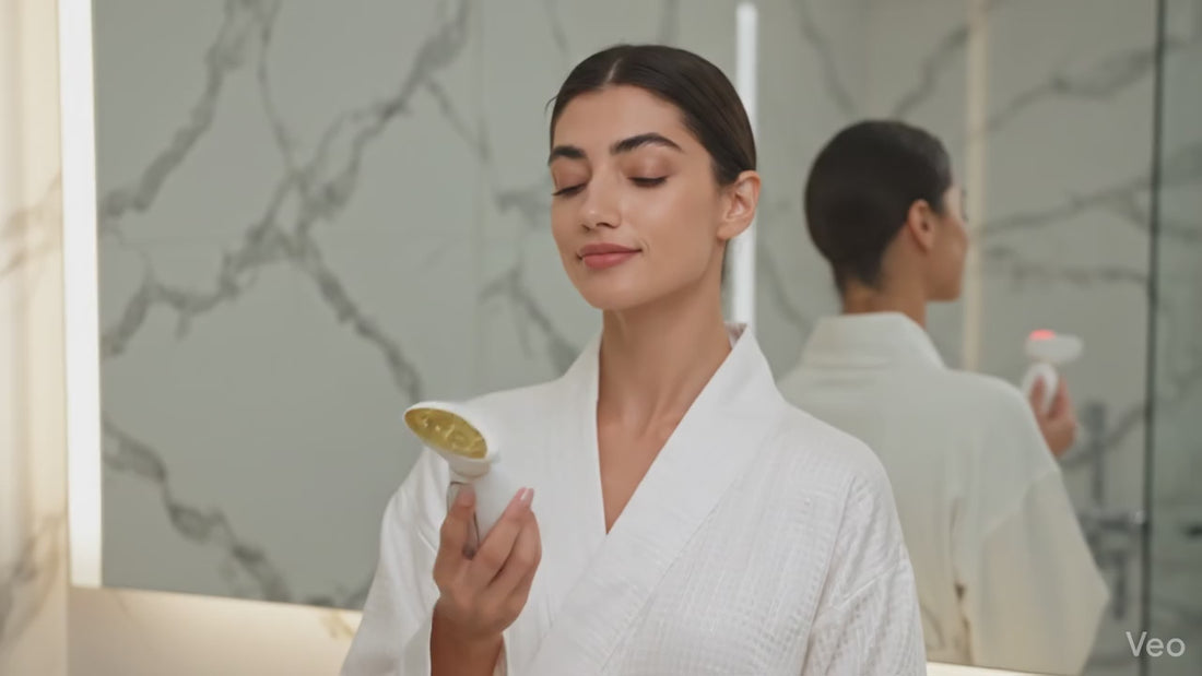 Démonstration utilisation appareil lifting visage PureBeam blanc et or. Femme massant son cou et son visage dans une salle de bain de luxe pour un effet raffermissant immédiat.